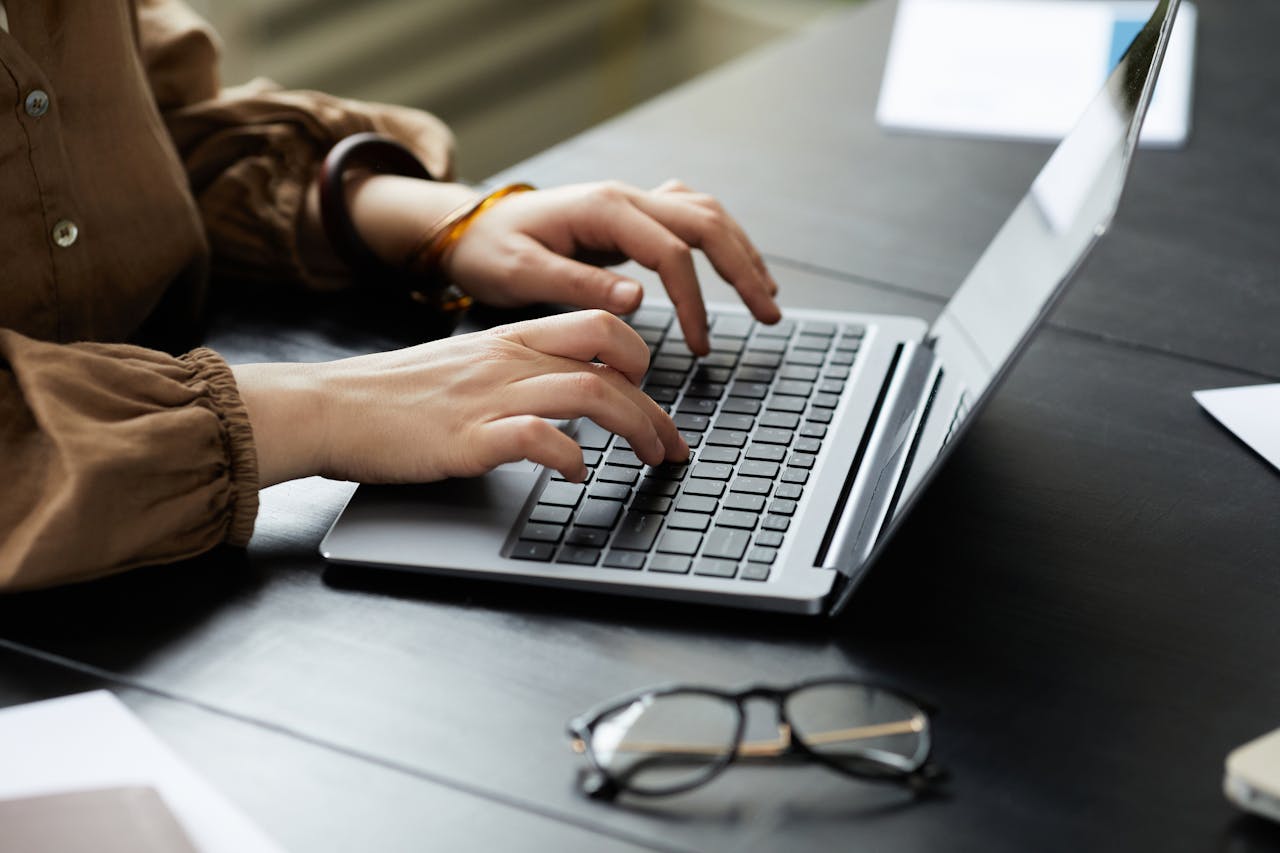 Hands typing on a laptop, eyeglasses on desk, modern workplace setting.